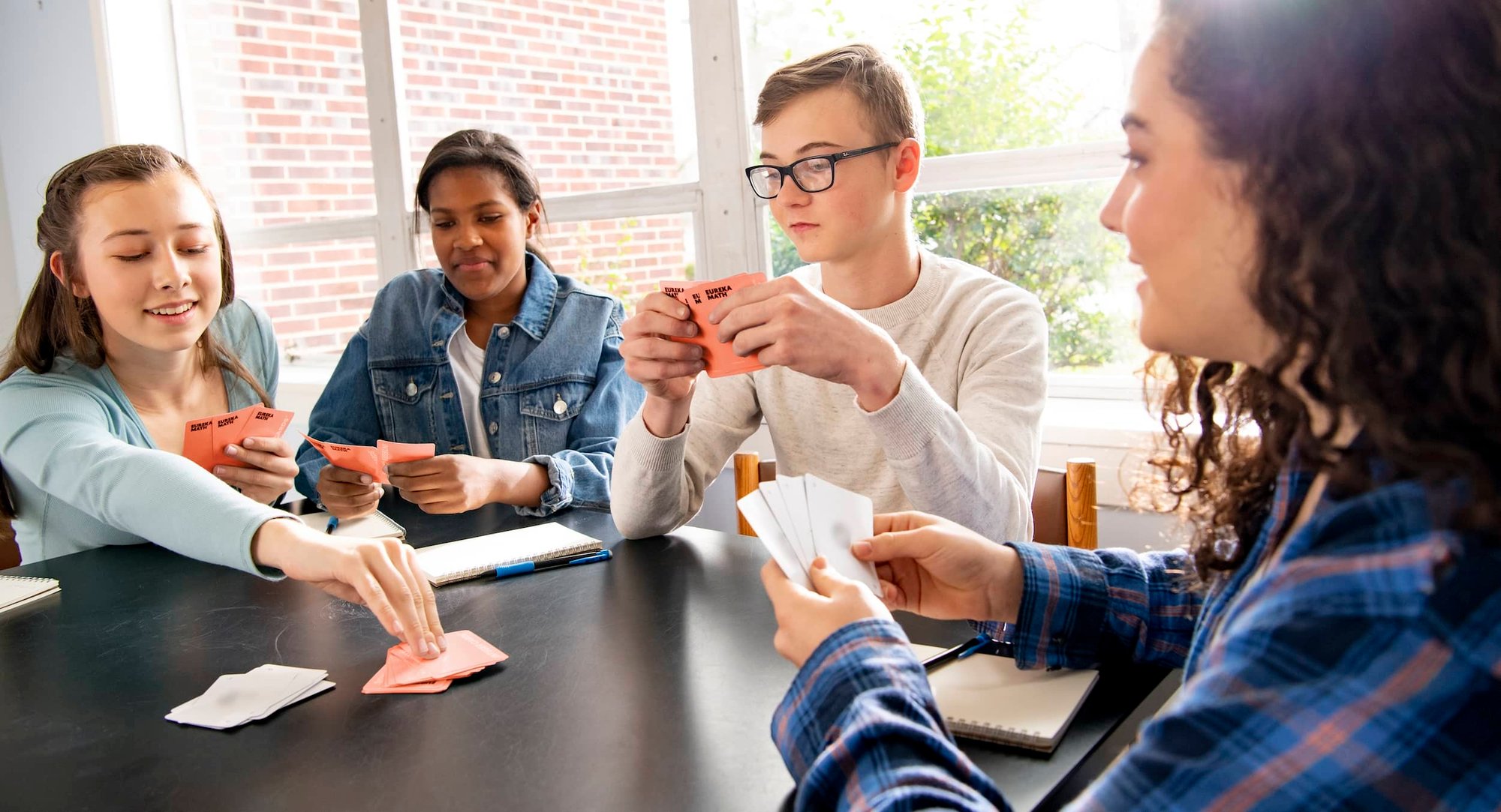 A group of four middle school age students sitting around a table. They are each holding some playing cards in their hands. There are piles of cards on the table in front of the students. One student has a pencil and notebook next to them.