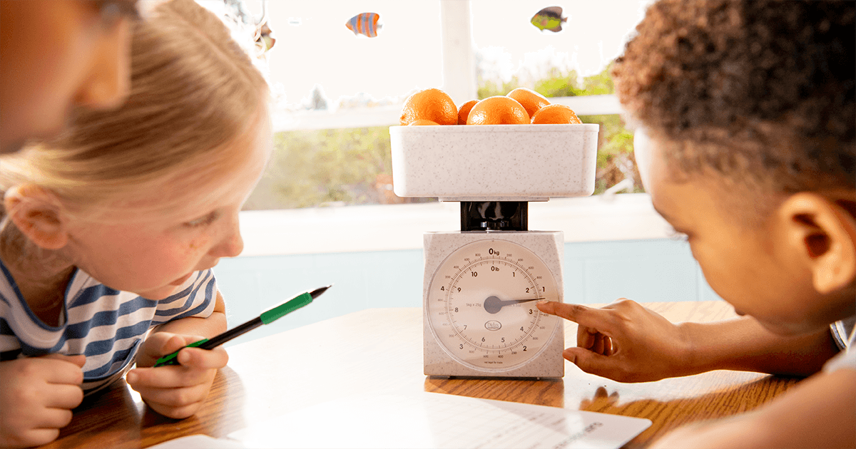 Three elementary students engage in a hands-on learning activity using a scale to weigh a bowl of oranges. One student points to the scale's dial while another takes notes with a pen, fostering collaboration and practical application of math concepts, as part of a Eureka Math² lesson.